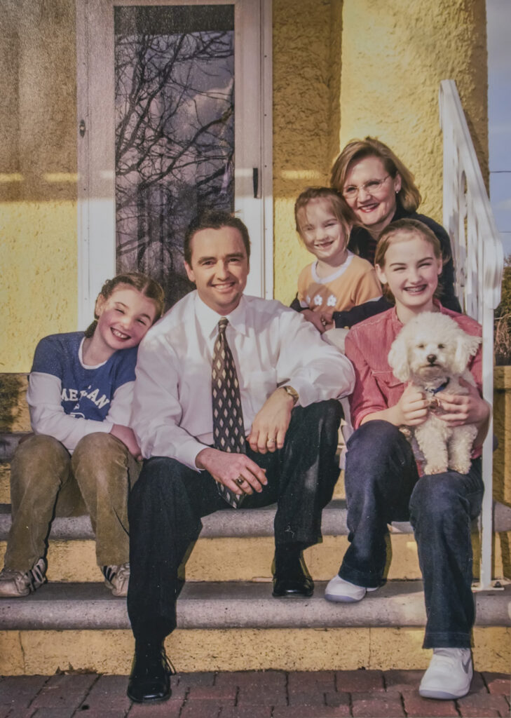 Jim and Dona Schmitt seated on their front steps with three daughters and a dog.