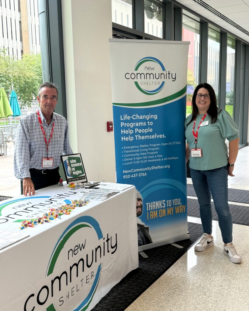 Two people stand beside a New Community Shelter information table and banner at an indoor workplace volunteer event.
