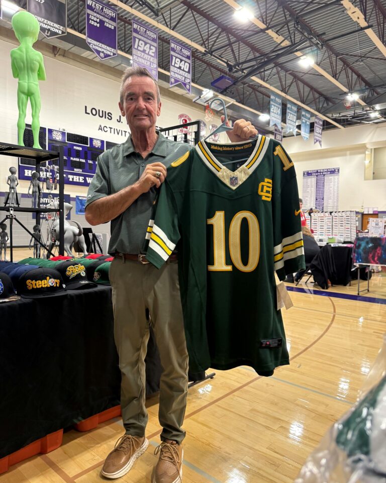 Jim Schmitt stands in the West High School gym holding an oversized green football jersey at a craft sale booth.