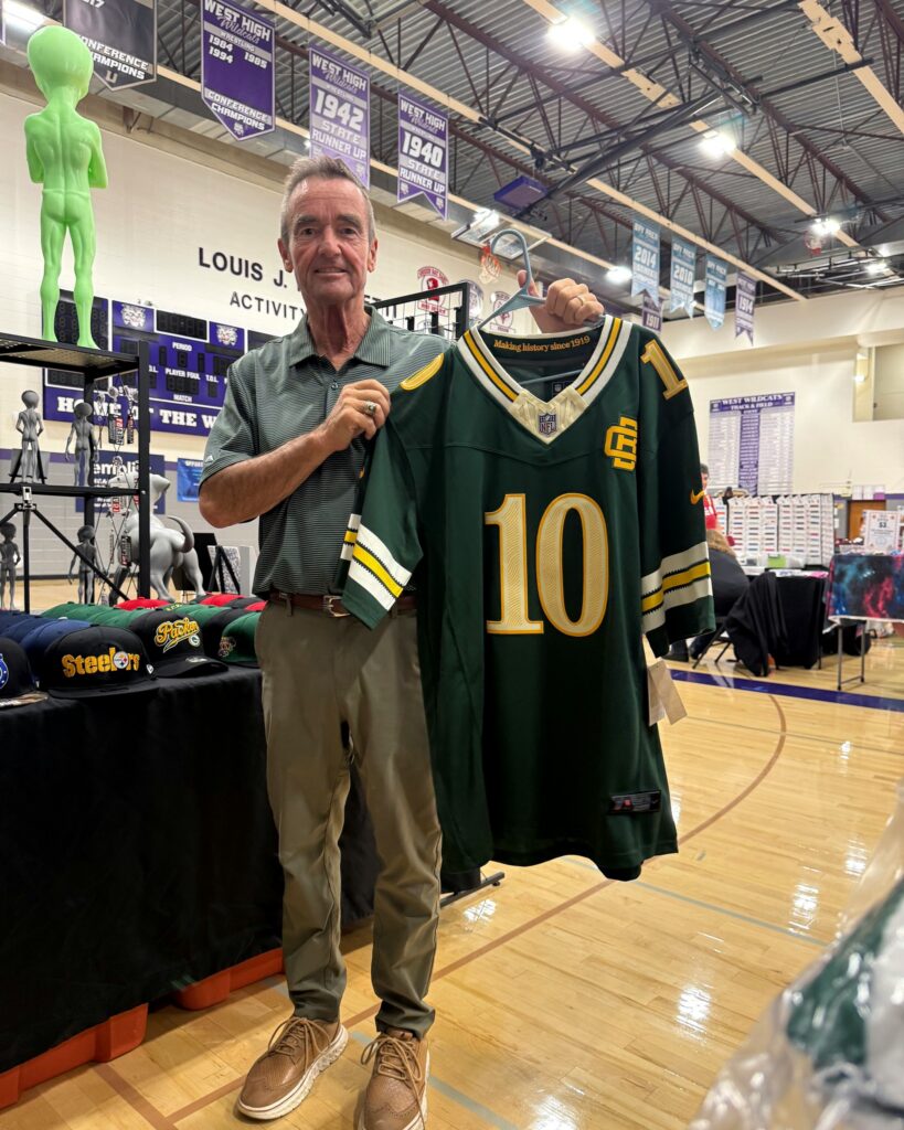 Jim Schmitt stands in the West High School gym holding an oversized green football jersey at a craft sale booth.
