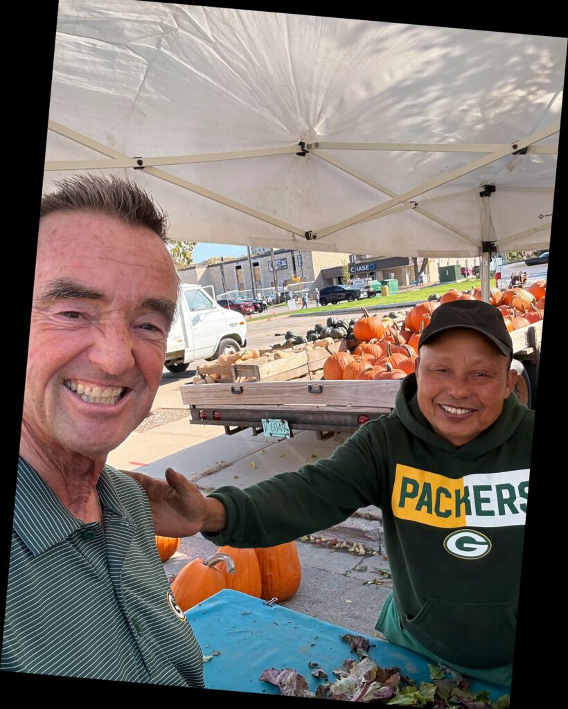 Jim Schmitt smiles with a vendor at a downtown farmers market stand with pumpkins and produce behind them.