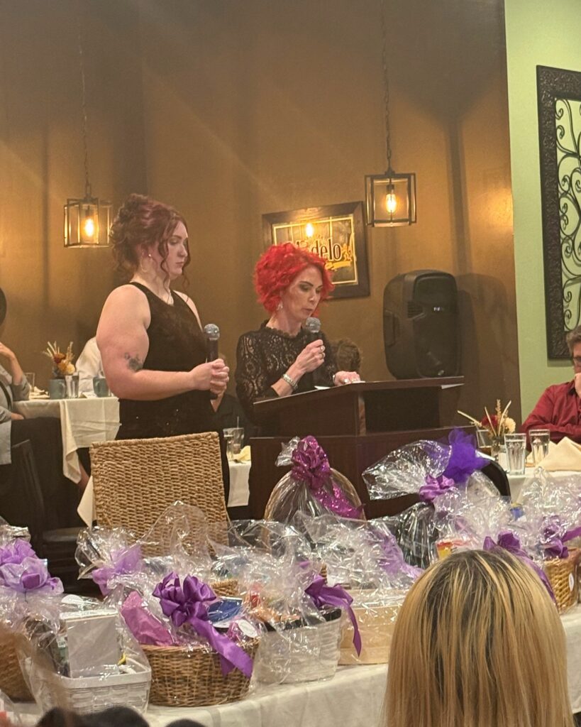 Two speakers hold microphones at a Serenity House of Green Bay gala fundraiser, with donated baskets on tables in the foreground.